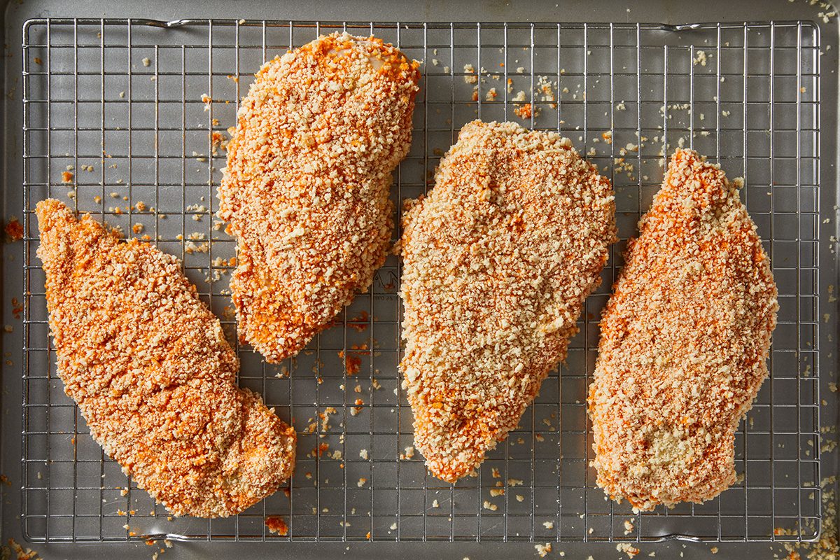 Four breaded chicken breasts are arranged on a wire rack set over a baking sheet, ready to be baked. The crumbs are golden and evenly coat the chicken pieces.