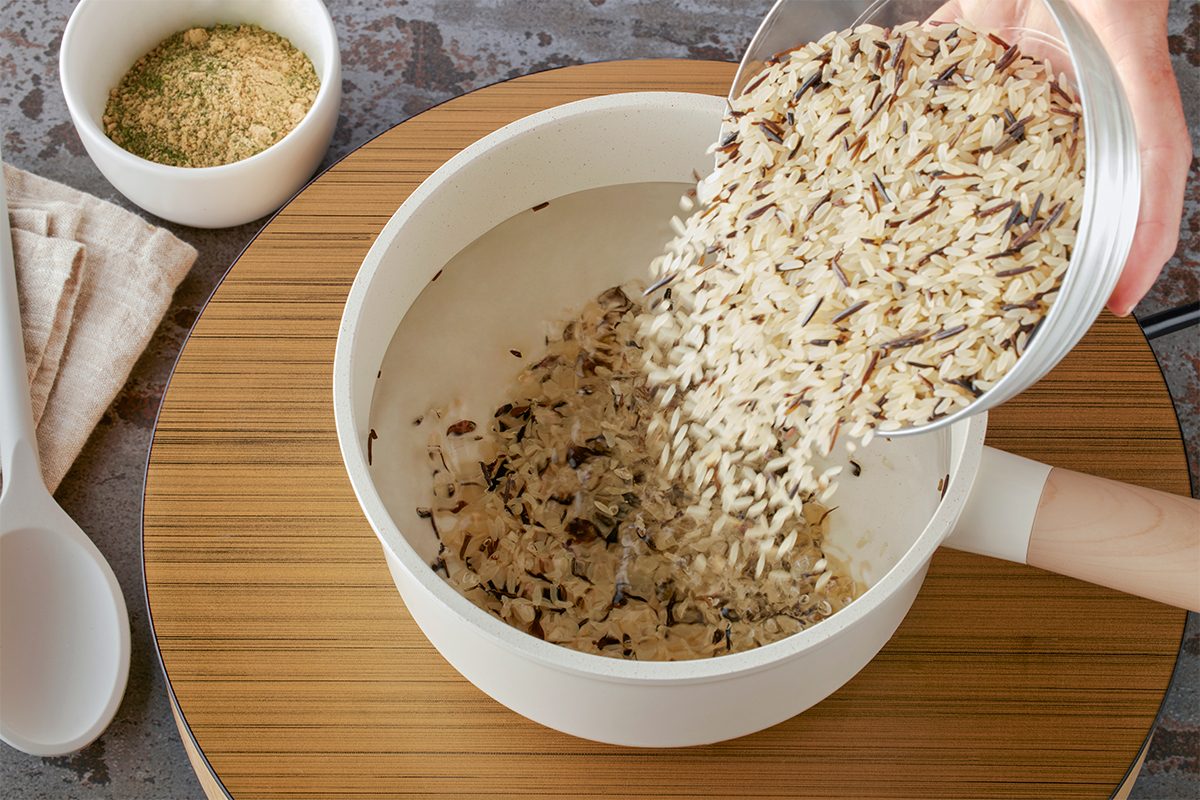 Overhead shot of a hand pouring uncooked wild rice blend into a white pot of boiling water on a wooden surface, with a white spoon, beige napkin, and a small bowl of seasoning nearby;