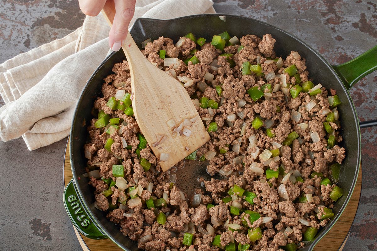 Overhead shot of a hand stirring ground beef, green bell pepper, and onions in a black skillet with a wooden spatula, with a beige towel resting on the counter nearby;