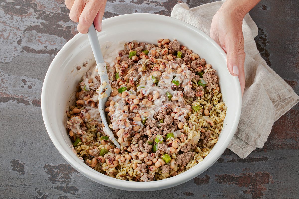 Overhead shot of someone mixing a casserole in a large white bowl using a white spoon while holding a light cloth, with beef, peas, rice, and peppers visible in the dish;