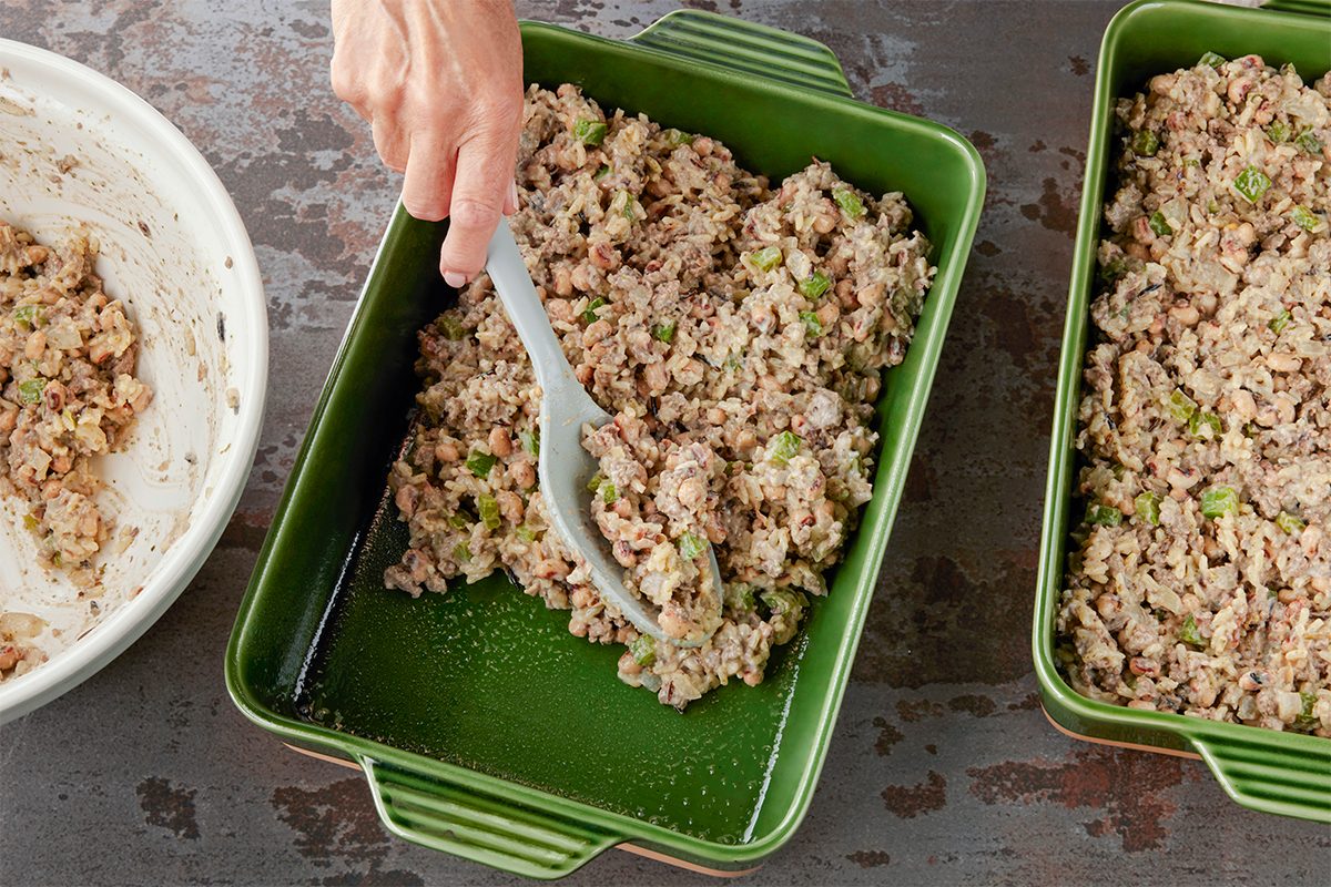 Overhead shot of a hand spreading a cooked mixture of rice, vegetables, and ground meat into a green rectangular baking dish, with more of the mixture in a bowl and another empty baking dish nearby;