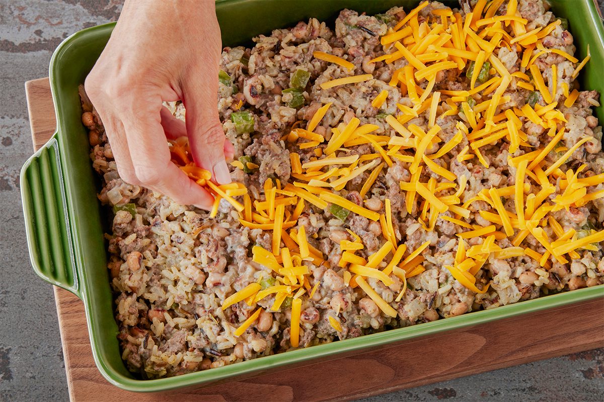 Overhead shot of a hand sprinkling cheddar cheese over a casserole in a green baking dish filled with rice, beans, and vegetables, placed on a wooden board;