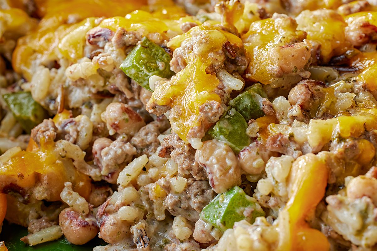 Closeup shot of Black-Eyed Pea Casserole, showing layers of ground meat, rice, and beans, topped with green bell peppers and gooey melted cheddar cheese;