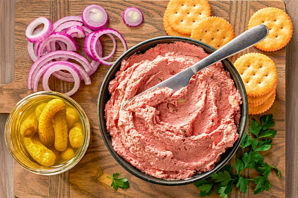 Overhead shot of a bowl of Braunschweiger spread with a spreading knife, surrounded by crackers, parsley, sliced red onions, and pickles arranged on a wooden board;