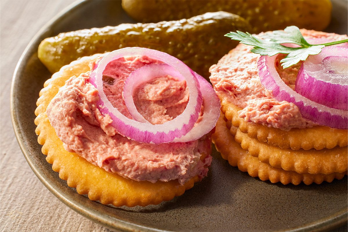 Closeup shot of Braunschweiger spread on round crackers topped with pink pâté, red onion rings, pickles, and a parsley sprig arranged on a plate