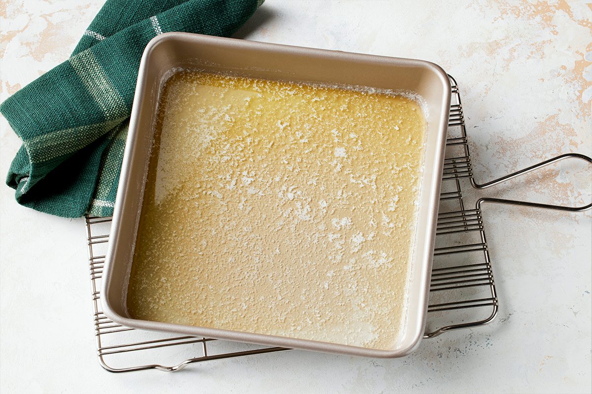 Overhead shot of a shiny square metal baking pan containing melted butter rests on a wire cooling rack beside it lies a green kitchen towel on a pale surface