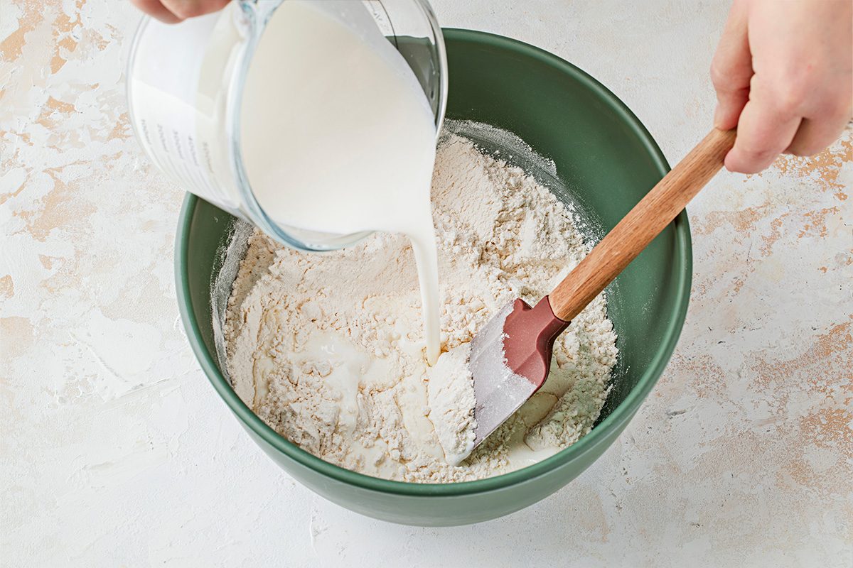 Overhead shot of a person holds a glass measuring cup of milk pouring it into a green bowl with flour and stirs with a bright red spatula on a plain surface