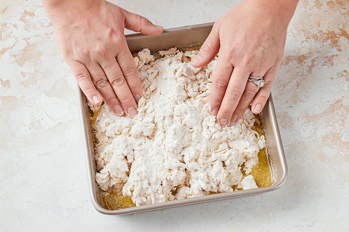 Overhead shot of a Two hands are pressing a crumbly dry dough mixture with visible flour and melted butter into a square metal baking pan on a pale surface