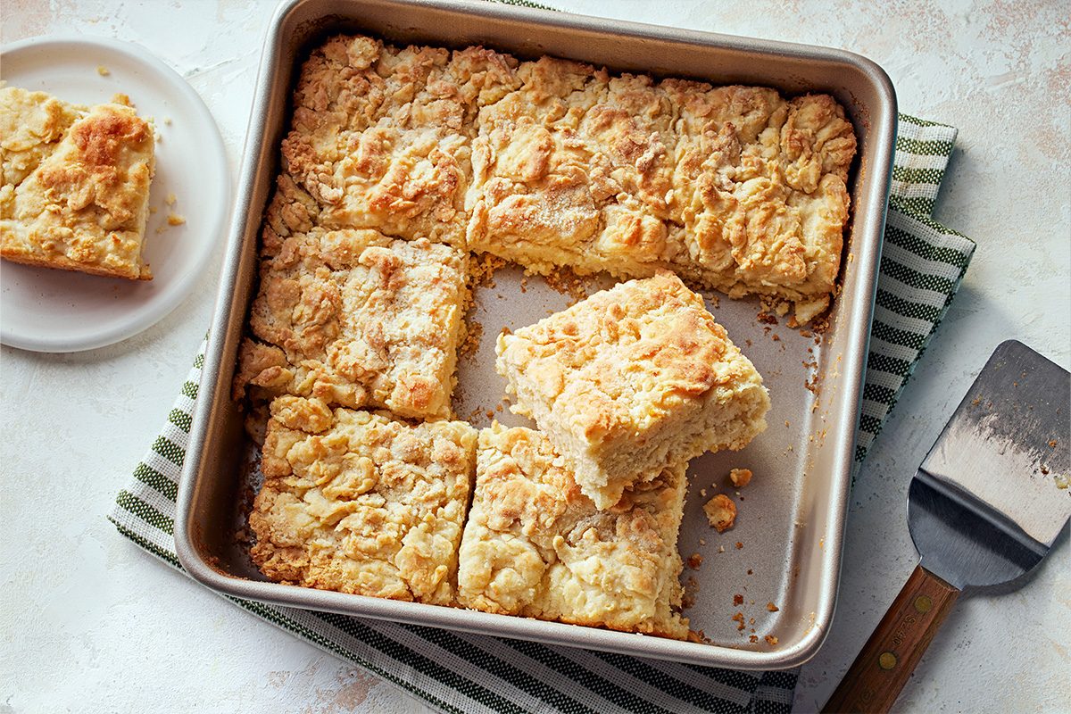 Overhead shot of a baking pan holds golden crumbly Butter Swim Biscuits some square and one lifted out a plate with another biscuit on a striped towel nearby a metal spatula rests next to the pan