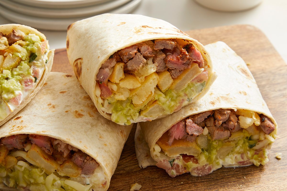 Three halves of burritos filled with steak, french fries, guacamole, and melted cheese are stacked on a wooden cutting board, with a plate visible in the background.