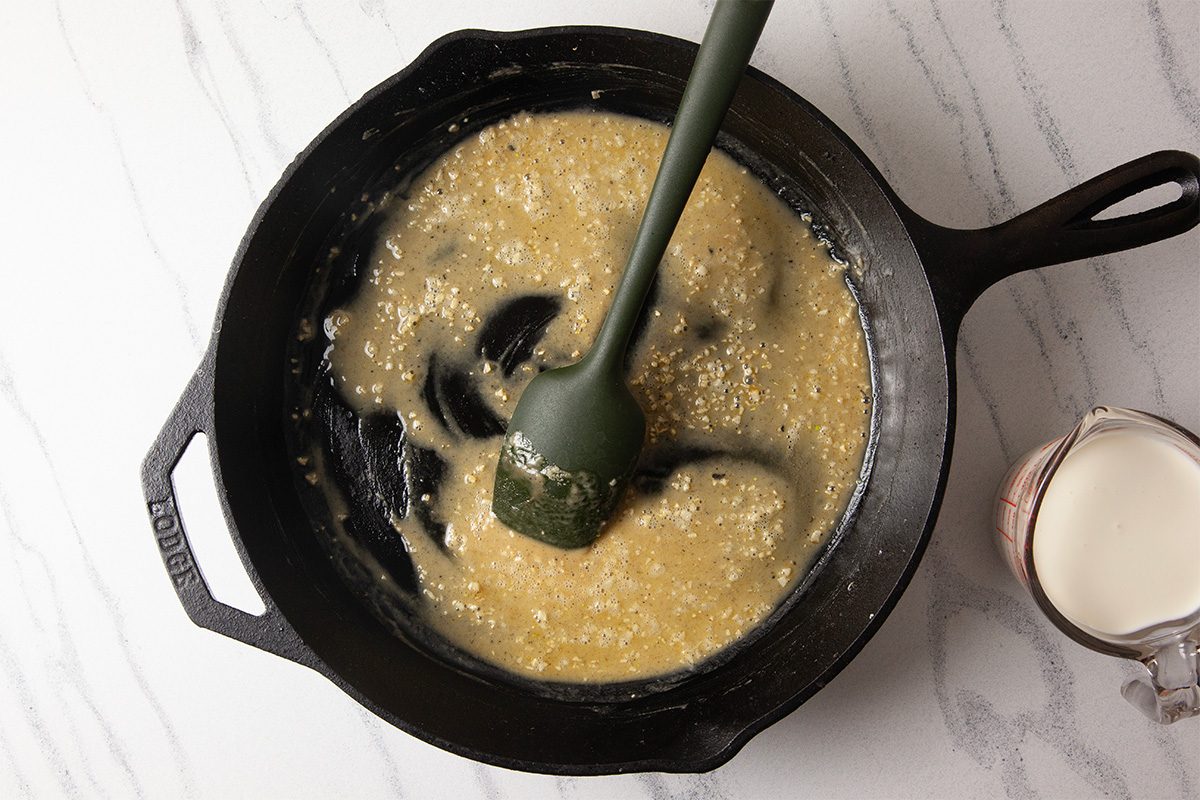 An overhead step-by-step cooking image showing butter, flour, and minced garlic being stirred together in a black cast-iron skillet to form the base of a Cheesy Chicken Alfredo Dip. The creamy roux is thickening as it cooks, demonstrating the first stage of preparing a rich Alfredo-style sauce.