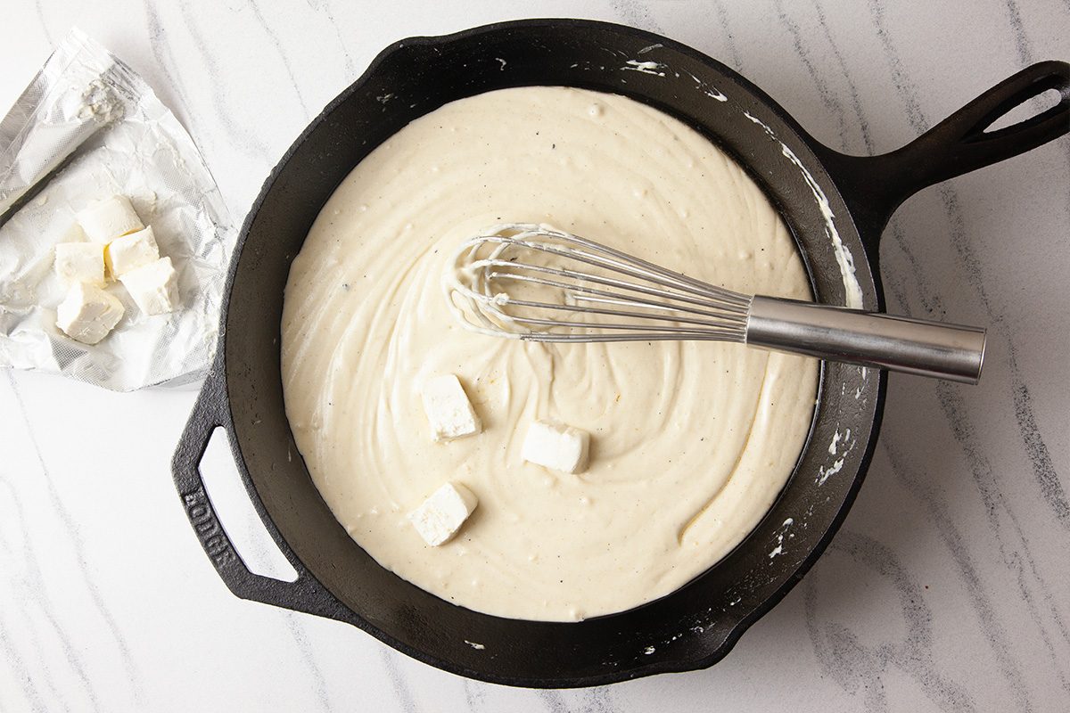 An overhead image of softened cream cheese and heavy whipping cream being whisked together in a cast-iron skillet to create a smooth Alfredo-style sauce. This step highlights the creamy foundation of Cheesy Chicken Alfredo Dip before seasoning and cheese are added.