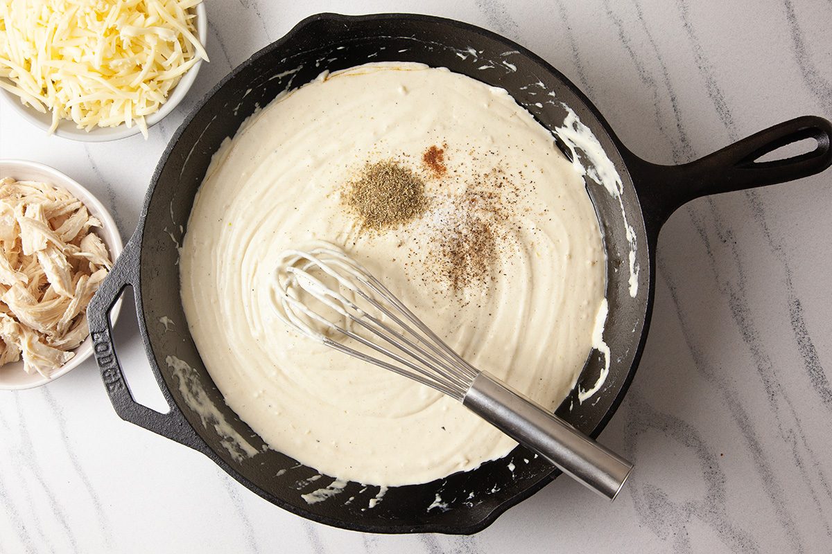 A top-down cooking image showing Italian seasoning, pepper, and freshly grated nutmeg sprinkled into a smooth Alfredo sauce in a cast-iron skillet. A whisk rests in the pan as the sauce is seasoned for Cheesy Chicken Alfredo Dip, emphasizing flavor development.