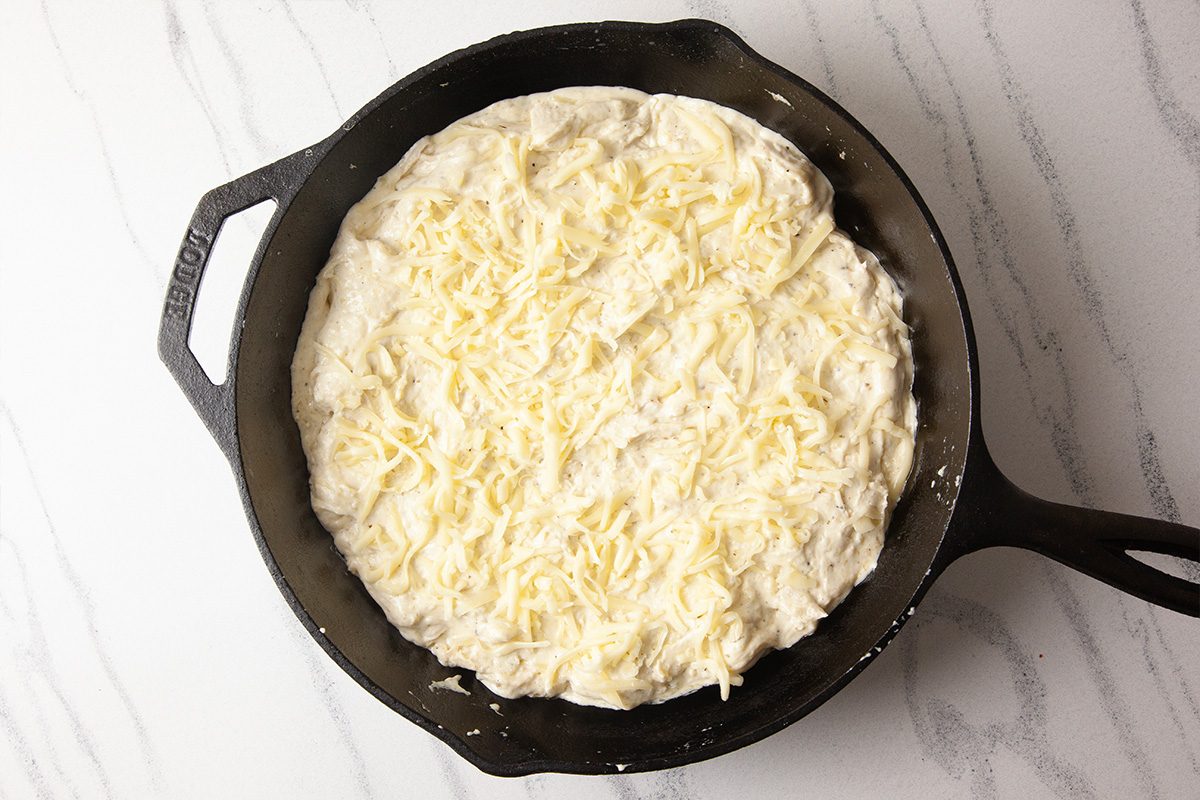 An overhead image of shredded mozzarella cheese evenly spread over creamy Alfredo sauce in a cast-iron skillet. This step shows Cheesy Chicken Alfredo Dip ready to be baked, highlighting the generous cheese topping before it melts.
