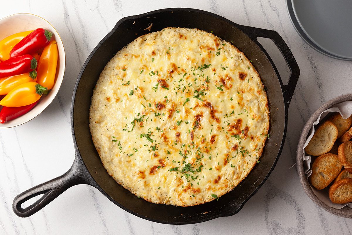 An overhead image of Cheesy Chicken Alfredo Dip baked until golden and bubbly in a cast-iron skillet. The melted mozzarella and Parmesan cheese are lightly browned and finished with minced fresh parsley, signaling the dip is ready to serve.