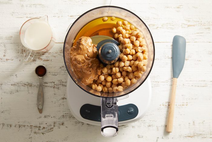 Top-down view of a food processor containing chickpeas, peanut butter, honey, and vanilla. Beside it are a rubber spatula, a spoon with cocoa powder, and a cup of milk on a white wooden surface.