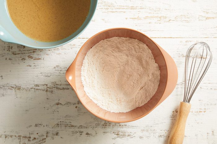 A bowl of flour, a whisk, and a mixing bowl with a tan batter sit on a white wooden surface, ready for baking.