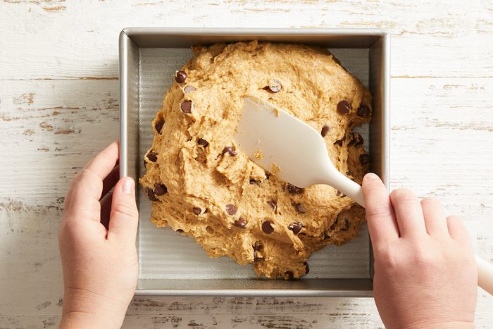 A person spreads chocolate chip cookie dough in a square metal baking pan with a white spatula on a light wooden surface.