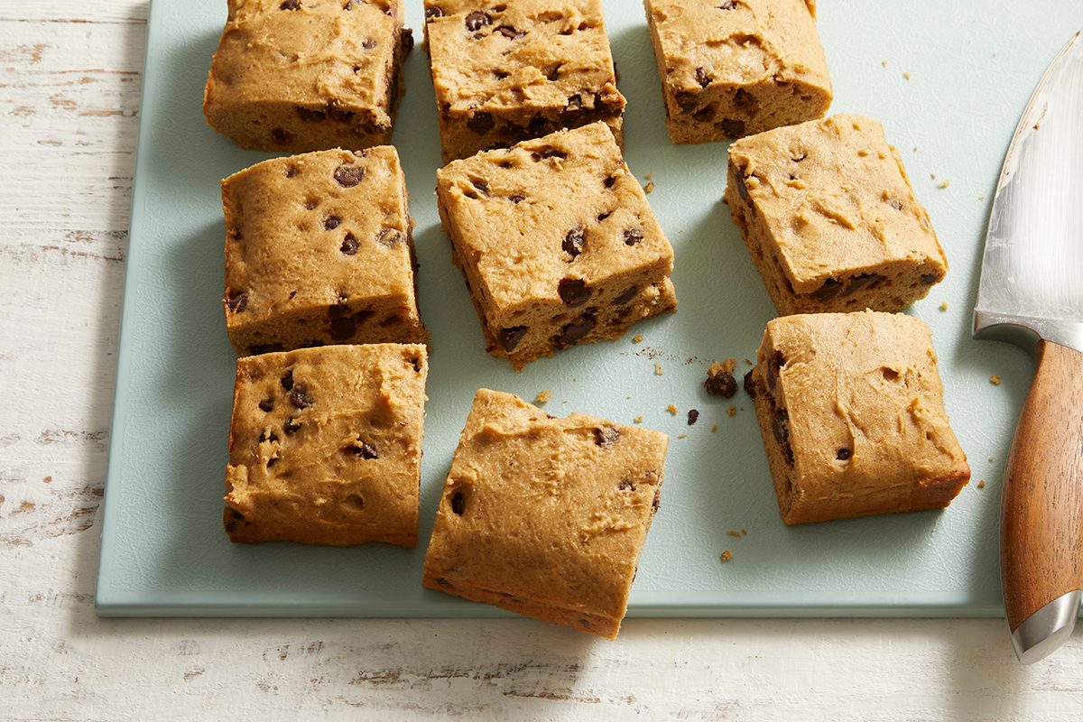 Nine square pieces of chocolate chip cookie dough are arranged on a light blue cutting board, with a large knife with a wooden handle resting nearby.