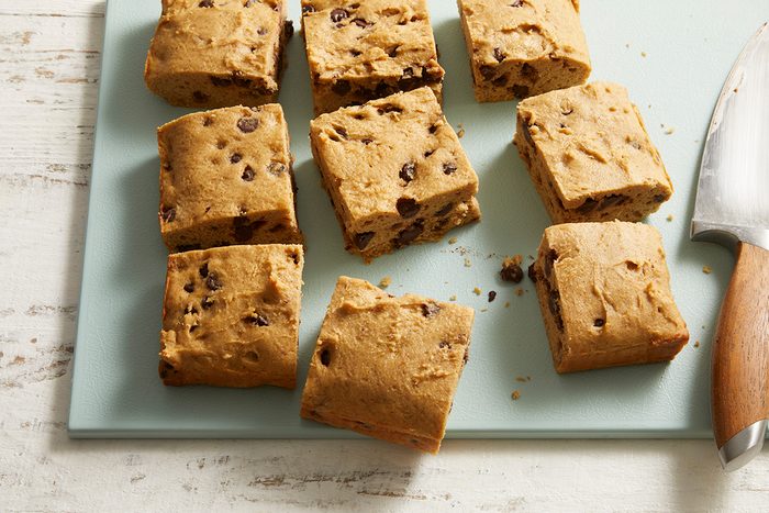 Nine square pieces of chocolate chip cookie dough are arranged on a light blue cutting board, with a large knife with a wooden handle resting nearby.