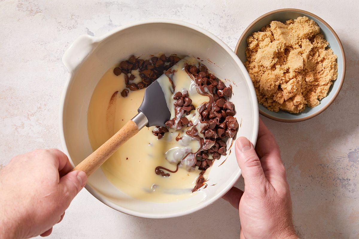 A person stirs chocolate chips and sweetened condensed milk in a white bowl with a spatula; a separate bowl of brown sugar sits nearby on a light surface.