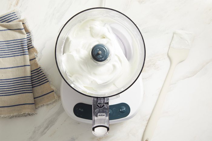 Overhead shot of a food processor filled with whipped, fluffy white cream on a light countertop, with a striped cloth and a white spatula with cream nearby;