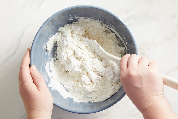 Overhead shot of a person stirring flour and other dry ingredients in a blue mixing bowl with a white spatula on a white countertop;