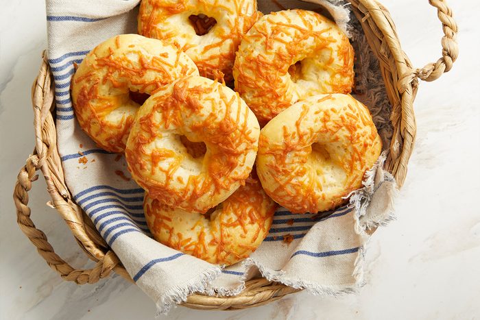 Overhead shot of golden, cheese-topped cottage cheese bagels arranged in a woven basket lined with a blue-striped cloth napkin on a light marble surface;