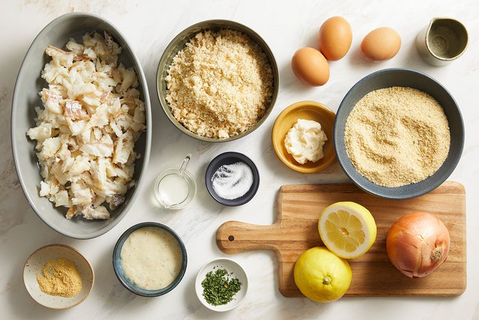 A variety of ingredients are arranged on a white surface, including chopped fish, breadcrumbs, eggs, mayo, onion, lemon halves, seasonings, and fresh herbs, some in bowls and some on a wooden cutting board.
