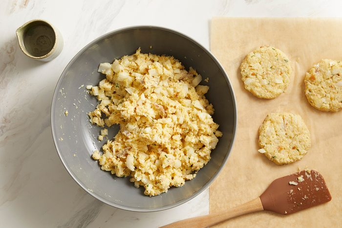 A gray bowl filled with a chopped, creamy egg salad mixture sits on a white surface next to three round scoops of salad on parchment paper and a wooden spatula, with a small cup nearby.