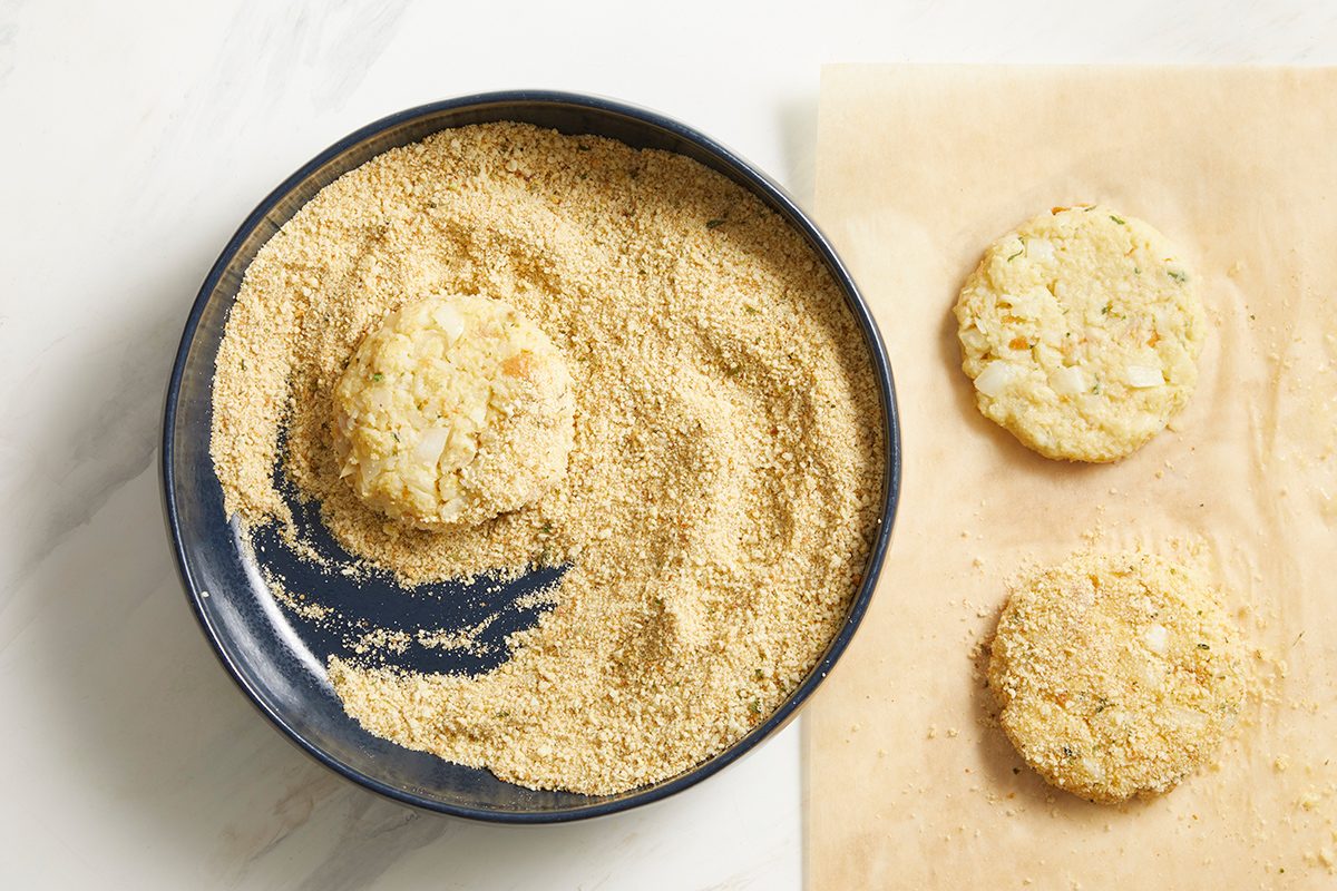 A round patty being coated in breadcrumbs in a dark bowl, with two breadcrumb-coated patties resting on parchment paper beside the bowl.