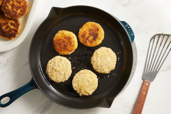 Five breaded patties are cooking in a black skillet; three are uncooked, and two are browned. A spatula rests nearby on a white surface, with more patties visible on a plate to the side.