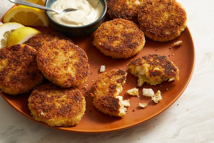 A plate of golden-brown crab cakes, some whole and one broken open, served with lemon wedges and a small bowl of creamy dipping sauce.