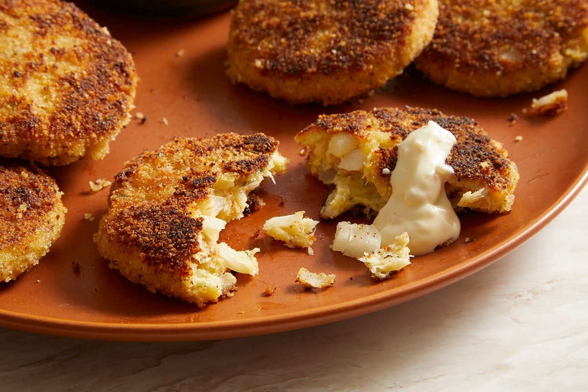 A brown plate with several golden-brown, breaded crab cakes. One crab cake is broken open, revealing the flaky interior, and topped with a dollop of creamy white sauce.