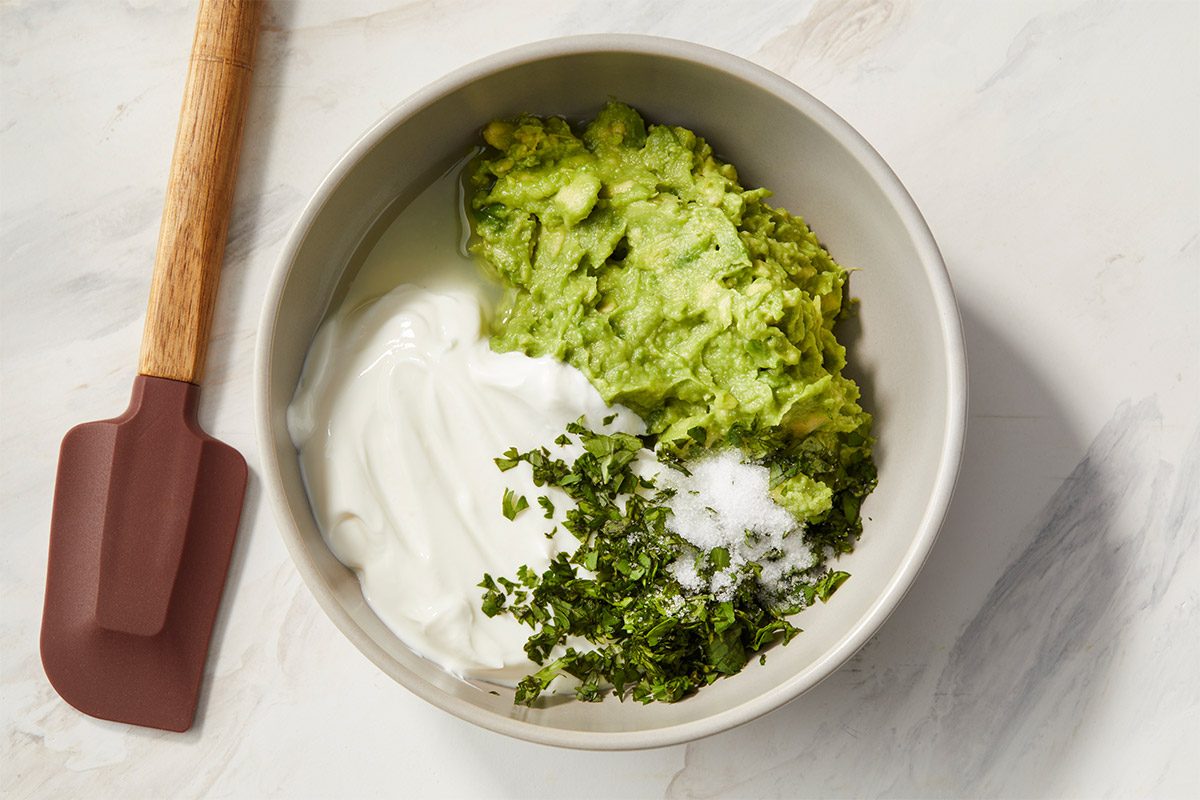 Bowl of mashed avocado, Greek yogurt, lime juice, fresh cilantro, and salt ready to be mixed into a smooth avocado crema. A spatula rests beside the bowl, showcasing preparation of a creamy sauce for a fish taco bowl.