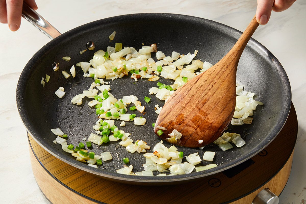 Chopped onion, garlic, and jalapeño peppers sauté in olive oil in a skillet, building flavor for the fish taco bowl base. A wooden spoon stirs the aromatic vegetables.