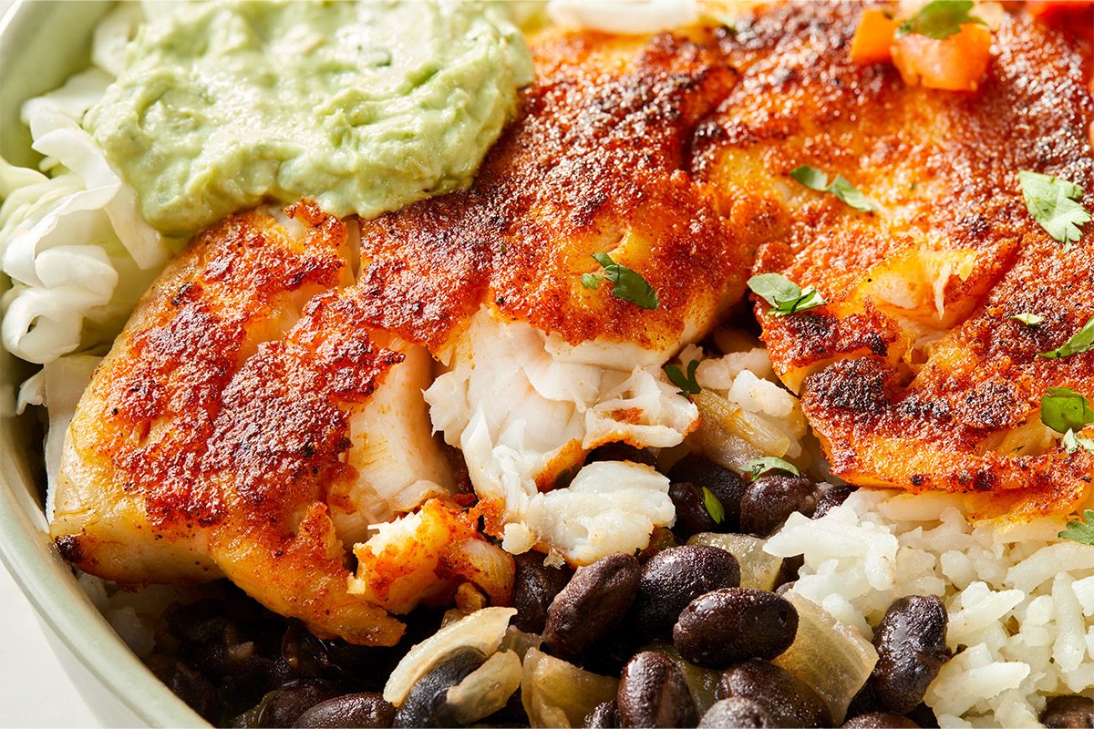 Close-up detail of the finished fish taco bowl highlighting flaky tilapia, seasoned black beans, cilantro rice, and creamy avocado crema. Vibrant textures and colors emphasize freshness and flavor.