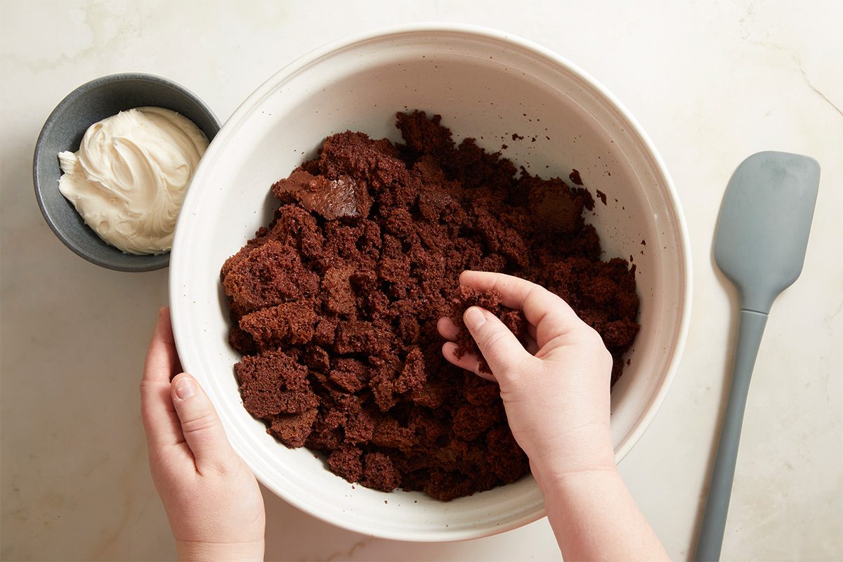 Overhead step-by-step food photography showing hands mixing crumbled chocolate cake in a white bowl with cream cheese frosting to form the base for football cake pops on a light neutral surface.