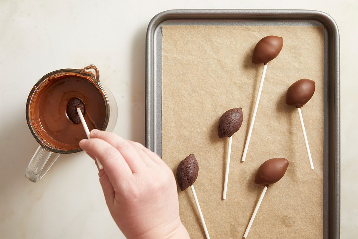 Overhead step-by-step image showing football-shaped cake pops being dipped into melted chocolate coating using lollipop sticks.