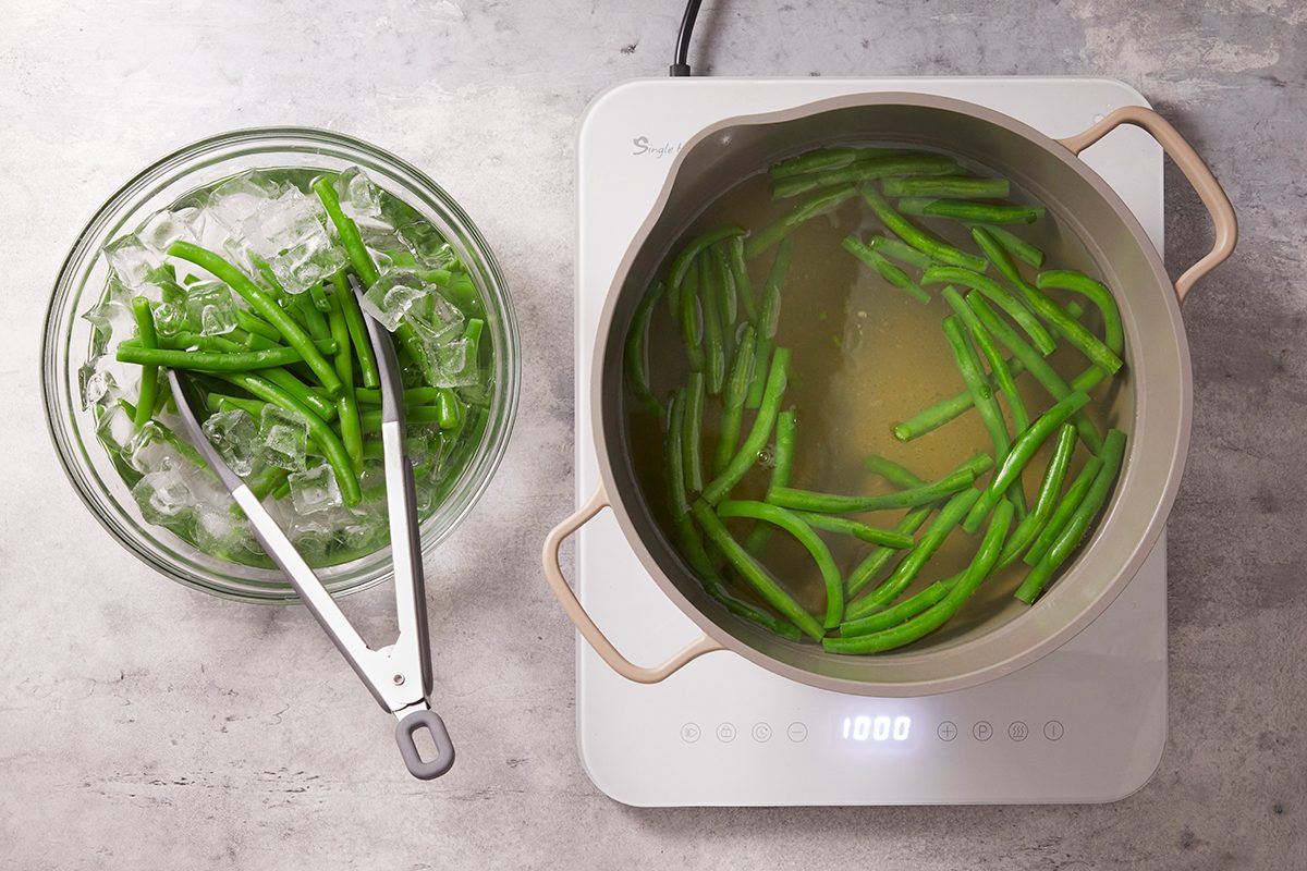 Green beans are being blanched on a stovetop; some are in a pot of boiling water while others are in a bowl of ice water with tongs resting on the bowl.