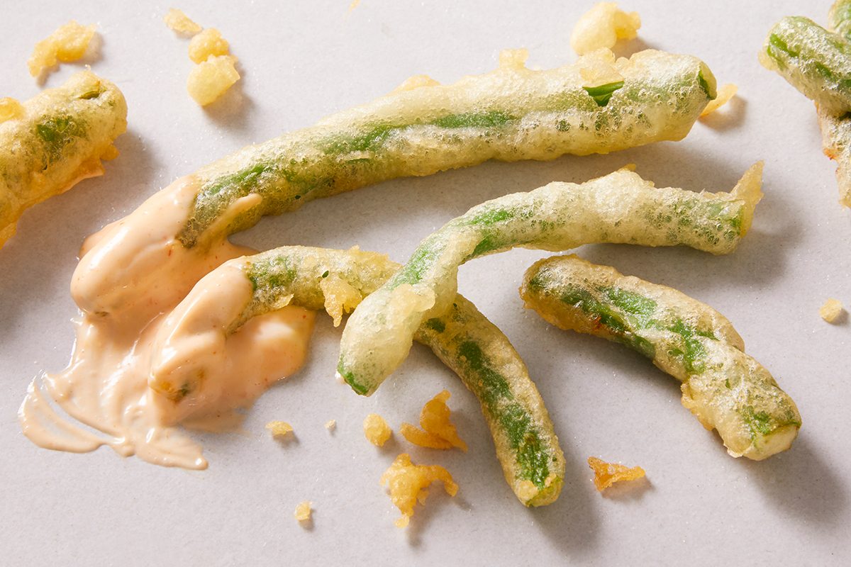 Crispy tempura green beans with a light golden batter are scattered on a white surface, with a small portion dipped in a creamy orange-colored sauce.