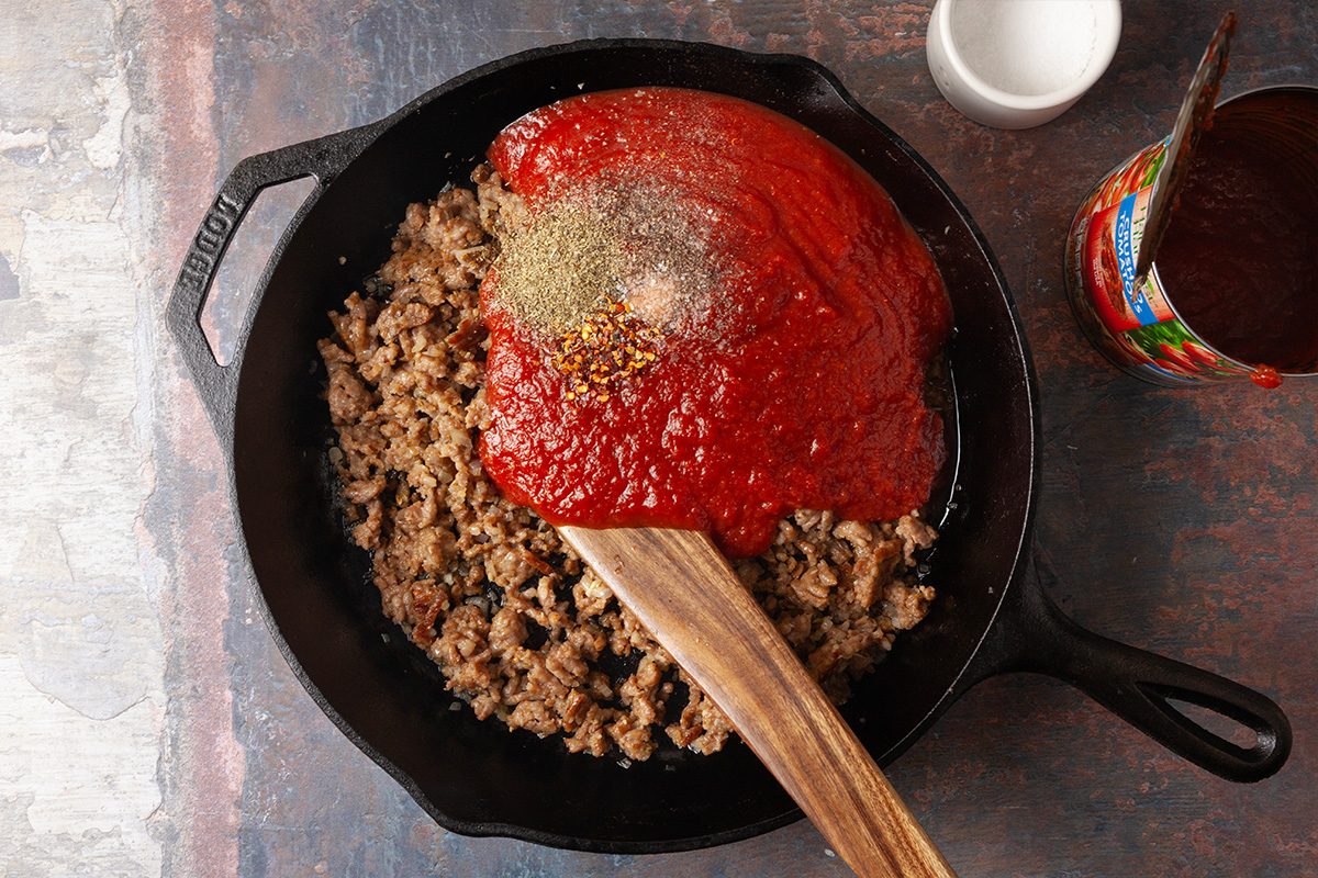 Overhead, horizontal step-by-step image of crushed tomatoes and seasonings being added to browned Italian sausage in a cast iron skillet. The tomato sauce begins forming the base of a gnocchi lasagna bake.