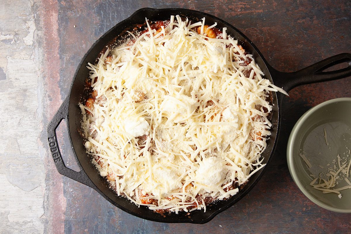 Overhead, horizontal step-by-step image of shredded mozzarella and Parmesan cheeses generously covering the gnocchi lasagna bake in a cast iron skillet before baking.