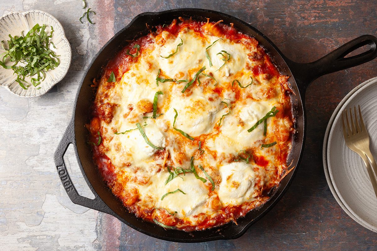 Overhead, horizontal image of a fully baked gnocchi lasagna with melted mozzarella, ricotta, and Parmesan cheese, finished with fresh basil in a cast iron skillet.