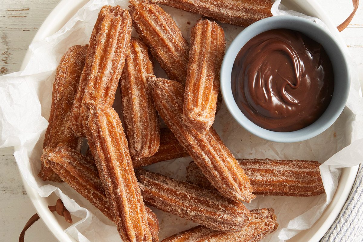 Overhead shot of Homemade Churros stacked in a white bowl lined with parchment paper, served with a small bowl of chocolate dipping sauce on the side and a dusting of cinnamon sugar.