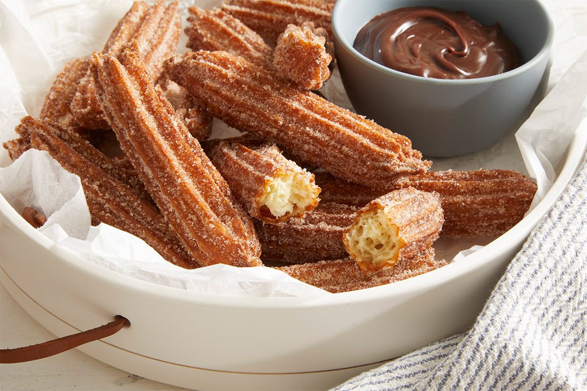 3/4 shot of Homemade Churros stacked in a white bowl lined with parchment paper, served with a small bowl of chocolate dip