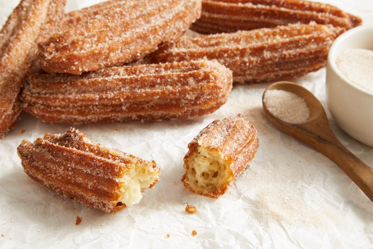 Close up shot of Churros arranged on parchment paper with a wooden spoon and a cup of chocolate sauce in the background.