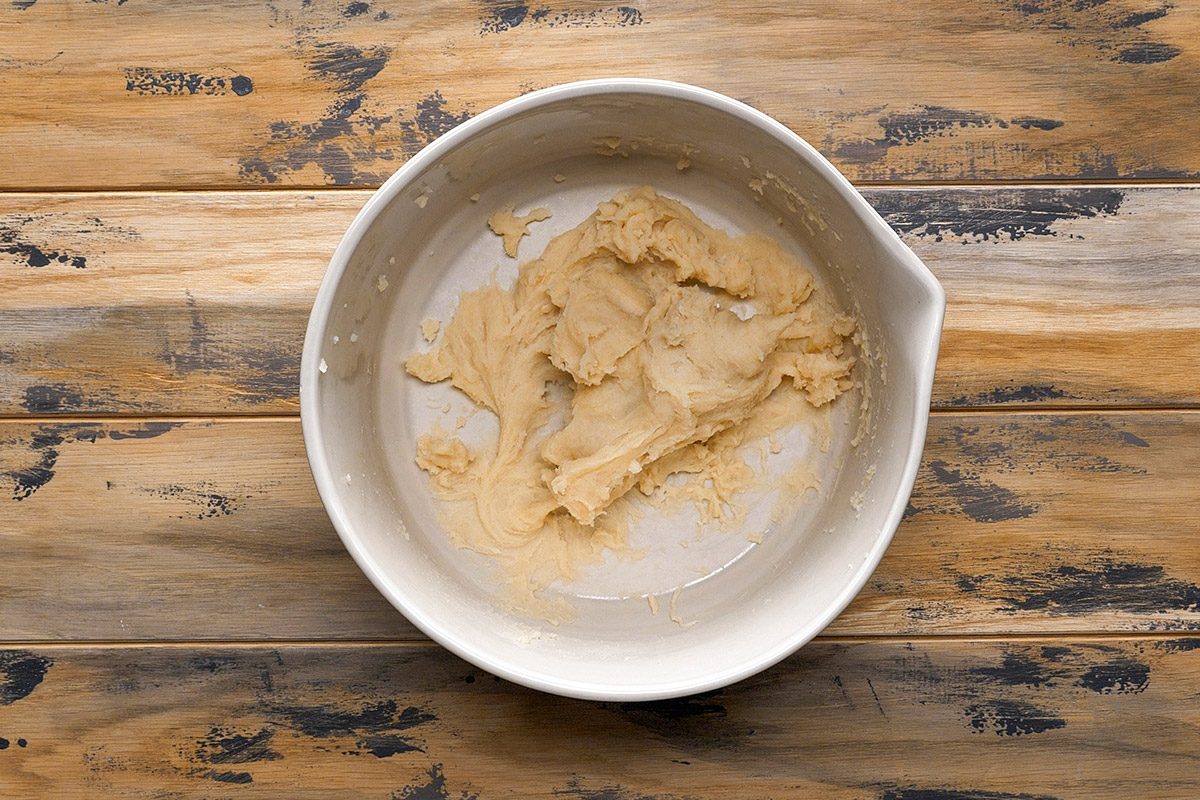 Overhead shot of a white mixing bowl holds thick cookie dough as it rests on a rustic wooden table and the surface shows a weathered and aged finish