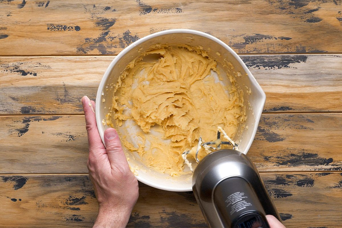 Overhead shot of a person is blending thick yellow batter in a white bowl using an electric hand mixer on a rustic wooden surface and mixing thoroughly
