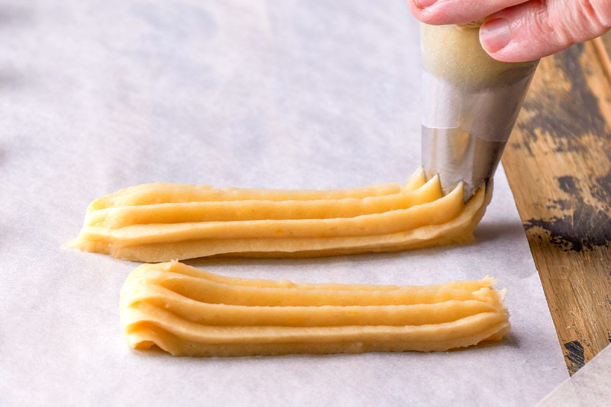 3/4th shot of a hand uses a piping bag with a star tip to pipe dough onto parchment on a wooden surface dough forms rectangles for pastries